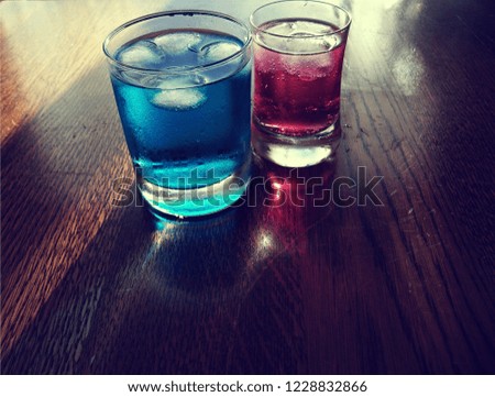2 refreshing cold blue and red soda drinks with lots of ice. Soda drinks are in small glasses. Soda drinks are transparent and some bubbles can be seen. 2 reflections can be seen on the wood.