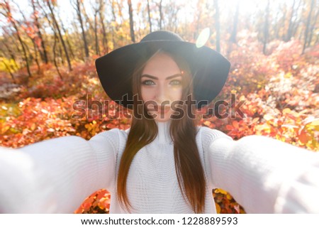 Portrait of beautiful young happy woman in black hat making selfie in the autumn park full of yellow leaves