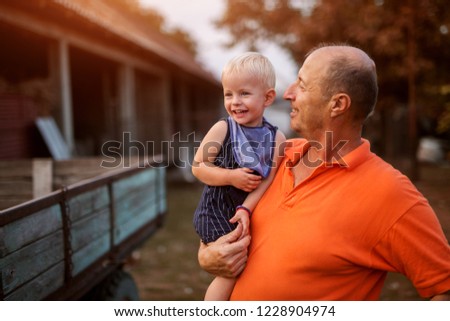 Picture of a proud grandfather holding his grandson and looking at him with love. Standing in a backyard of a farm.