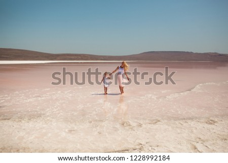 happy mom and daughter go by the hand to the pink lake, dressed in bright colors, mom has blond long hair