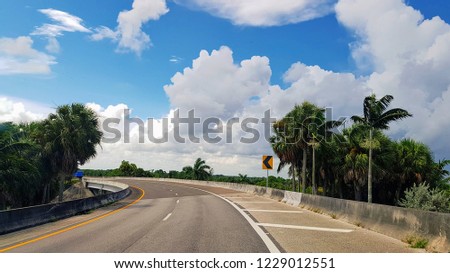 Summer travel in Florida by car, long empty highway road turning left, yellow road sign on the right, tropical palms on both sides of road, sunny sky with beautiful clouds in the background