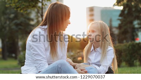Happy mother with her daughter are sitting in the park on the background of a beautiful sunset. Cute woman with a blonde daughter touch their noses and laugh.