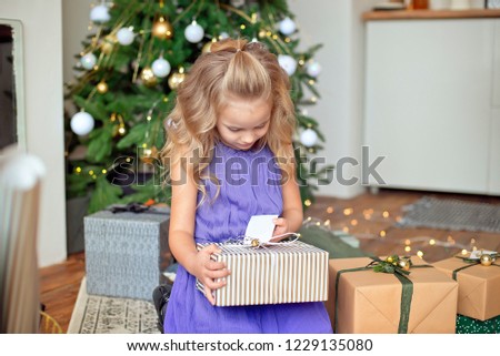 Little beautiful girl with blond curly hair stretches a Christmas gift against the background of the Christmas tree. Christmas gift, New Year's gift Christmas concept.