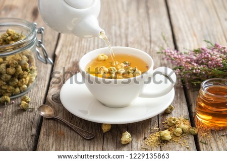 Healthy chamomile tea poured into white cup. Teapot, small honey jar,  heather bunch and glass jar of daisy medicinal herbs. Royalty-Free Stock Photo #1229135863