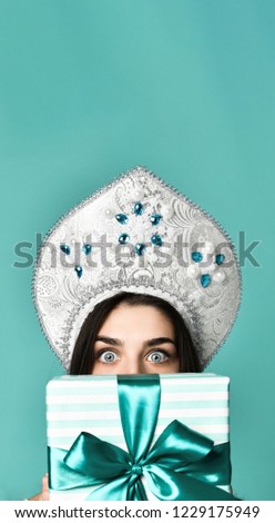 Portrait of a pretty smiling gir in traditional russian cap kokoshnik holding gift box and hiding behind him isolated over turquoise background