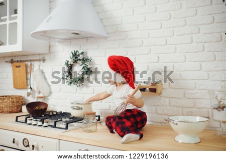 Cute little girl in santa hat, preparing cookies in the kitchen at home. Sits on the kitchen table and helps mom prepare a festive Christmas dinner