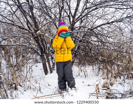 The child dabbles and plays outside in the winter. White snow background. Warm rainbow knitted hat and yellow jacket. Little boy