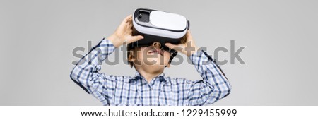 A charming boy with an inlaid shirt and light jeans stands on a gray background. The boy on his face glasses virtual reality