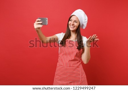 Housewife female chef cook or baker in striped apron, white t-shirt, toque chefs hat isolated on red wall background. Smiling fun woman doing selfie shot on mobile phone. Mock up copy space concept