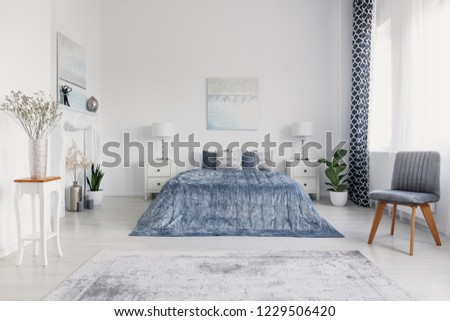 Grey chair next to blue bed in white bedroom interior with posters and lamps on cabinets. Real photo