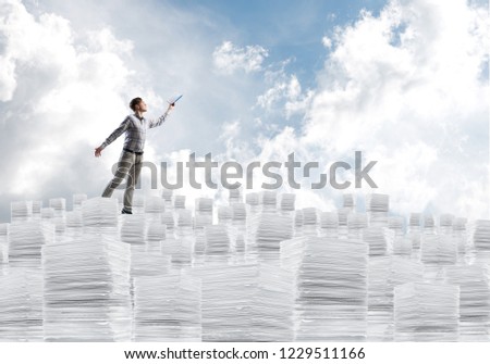 Man in casual wear keeping hand with book up while standing on pile of paper documents with cloudly skyscape on background. Mixed media.
