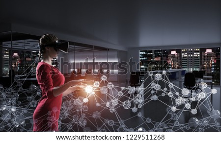 Horizontal shot of young and beautiful woman in red dress using virtual reality headset while standing indoors of dark office building with digital network structure