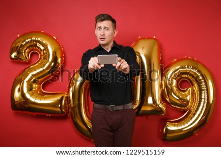 Merry fun young man in black shirt celebrating holiday party hold cellphone isolated on bright red wall background, golden numbers air balloons studio portrait. Happy New Year 2019 Christmas concept
