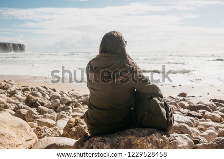 A girl with a backpack or a tourist or a traveler in solitude admires a beautiful view of the Atlantic Ocean in Portugal. Search for soul or unity with nature. Royalty-Free Stock Photo #1229528458