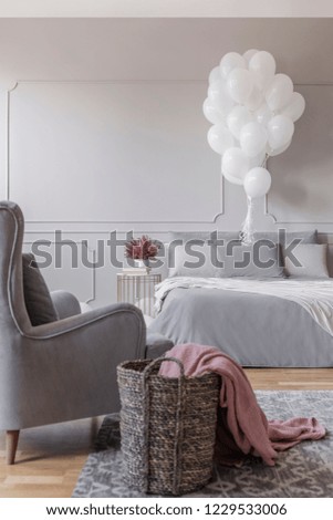 Birthday decoration in grey stylish bedroom with comfortable bed, trendy armchair and basket with pink blanket, real photo with copy space on the empty wall