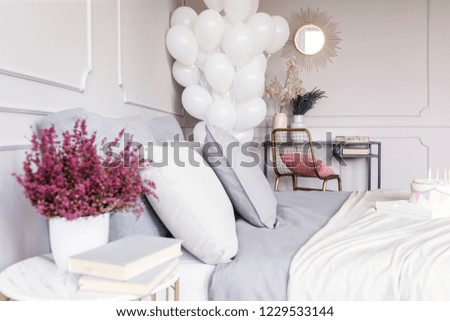 Closeup of heather and books on nightstand in cozy grey bedroom interior with double bed, birthday balloons and dresser with chair and mirror, real photo