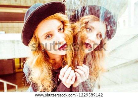 Beautiful young curly red hair woman with comedian black hat and her reflection in industrial stainless steel mirror surface wall. Strong and gentle, dual personality or alter ego psychology concepts