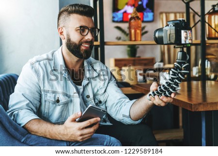 Young bearded male video blogger creates video content for his channel. Happy guy shoots video streaming for users while sitting in coffee shop. Man vlogger relieves himself on camera with tripod.