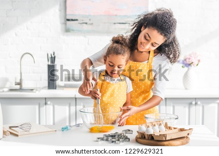 smiling african american mother helping daughter mixing eggs for dough in kitchen Royalty-Free Stock Photo #1229629321