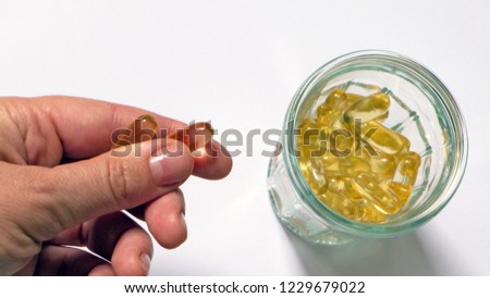 hand with two pills of fish oil on a leafy background with a glass jar next to it