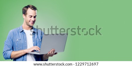 Smiling young man in blue smart casual wear, working with laptop, with empty copyspace area for slogan, advertisement or text message, over green background. Banner composition.