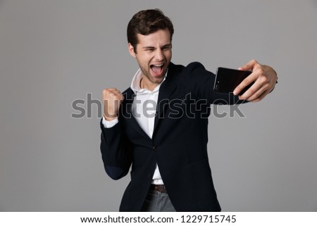 Image of cheerful man 30s in formal suit clenching fists while taking selfie photo on cell phone isolated over gray background