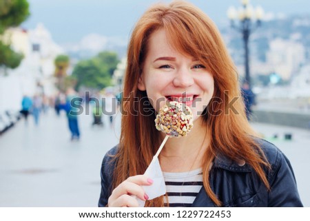 A nice, smiling, redhead girl is eating an apple in caramel in the city Royalty-Free Stock Photo #1229722453