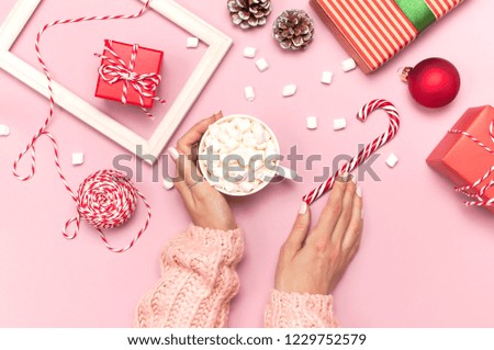 Female hands in knitted sweater holding cup of marshmallows and Christmas candy cane gifts boxes red ball photo frame on pink background top view Flat Lay. Winter traditional drink food. Festive decor