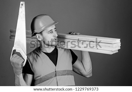 Woodcraft concept. Carpenter, woodworker, labourer, builder on thoughtful face carries wooden beams on shoulder. Man, handyman in helmet, hard hat holds handsaw and wooden beams, grey background.
