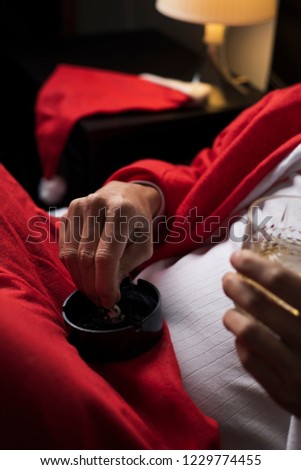 closeup of a young man wearing a santa suit drinking whisky and putting out a cigarette in an ashtray in bed