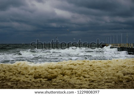 Sea storm with waves splashing at the pier at the beach with dark cloudy sky over it. Foam on the empty coastline.