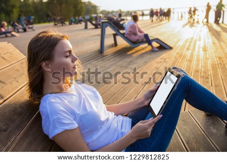 Attractive woman reading news in internet via modern touch pad with empty mock up copy space screen background for promotional content while sitting on bench  in park in sunny summer evening. 