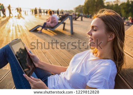 Smiling woman online booking via portable touch pad while relaxing outdoors in sunny summer evening during recreation time in vacation. Female reading e-book via digital tablet during rest 