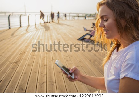Happy smiling hipster girl using messenger on mobile phone during leisure time in the fresh air.Female reading text message on cellphone, sitting on wooden quay with copy space for promotional content
