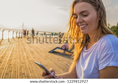 Happy smiling hipster girl watching video in social network via smartphone while sitting outdoors near copy space for promotional content. Cheerful female sending sms via cellphone during walking time