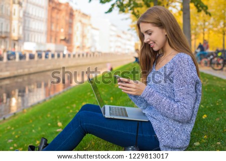 Attractive woman with smile on the face reading pleasant text message on mobile phone while sitting with laptop computer outdoors in park near embankment river background with copy space 