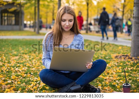 Young Caucasian female in stylish wear clever university student using laptop computer for preparing course work while siting on a campus in warm beautiful autumn day during free time after classes. 