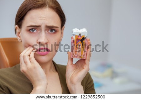 Young woman suffering from toothache, looking to the camera in despair, holding tooth mold showing cavity. Close up of a female patient with aching teeth visiting dental clinic. Dentistry, oral care Royalty-Free Stock Photo #1229852500
