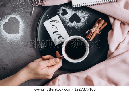 Valentines day background. Hand holds cup of coffee and notebook,  scarf, cinnamon and heart on gray table. Valentines Day concept. Flat lay, top view, copy space