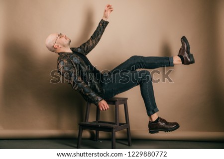 Indoor portrait of artistic adult unusual male. Funny bald man in biker leather jacket and oxford shoes. Fashion clothes. Stylish weird boy falling down from chair on brown background. Doing exercices