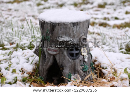 First snowfall of the year covers an outdoor decoration that looks like a tree stump with windows, a Welcome sign, and an opening as a Woodland animal home. The home and ground is covered with snow.