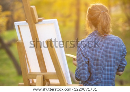 young woman artist standing near an easel and looking at the sunset on the lawn, the girl is looking for inspiration for painting a picture, a concept of hobbies and creativity