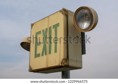 Old outdoor weathered ‘Exit’ sign with two lamps on the pole against cloudy sky background