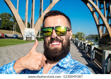 Everything is OK. Close up portrait of cheerful bearded guy. He is looking camera, showing thumb up sign and taking selfie with arch bridge on background