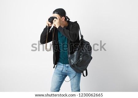 Guy with a black backpack on his shoulder dressed in a dark t-shirt, jeans, sweatshirt and a cap makes a photos in the studio on a white background