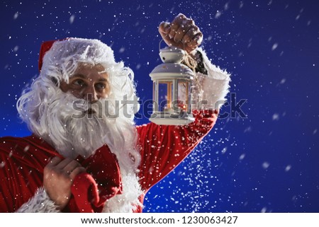 Santa Claus with a long white beard holds a lamp with a candle against a blue sky. It is snowing. Christmas