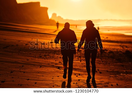Two people walking down the Algarve beach in Portugal during early morning backlit by the sun. 