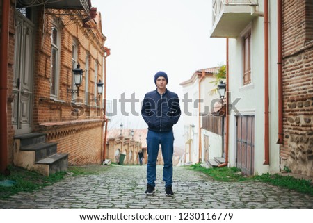 A handsome guy stands in front of old buildings. A handsome young man walks through the ancient streets. The guy walks through the cobbled streets. Royalty-Free Stock Photo #1230116779