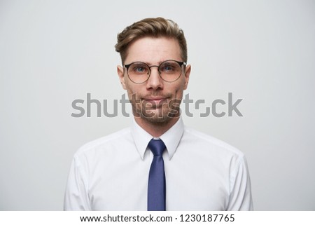 Photo as on passport. Young man with stylish haircut, glasses,blue eyes look straight little smiling and self-collected, dressed in white shirt and blue tie, unshaved, over white background