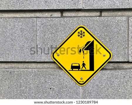 yellow sign on cement block wall, with warning illustration of snow falling from roof onto people 